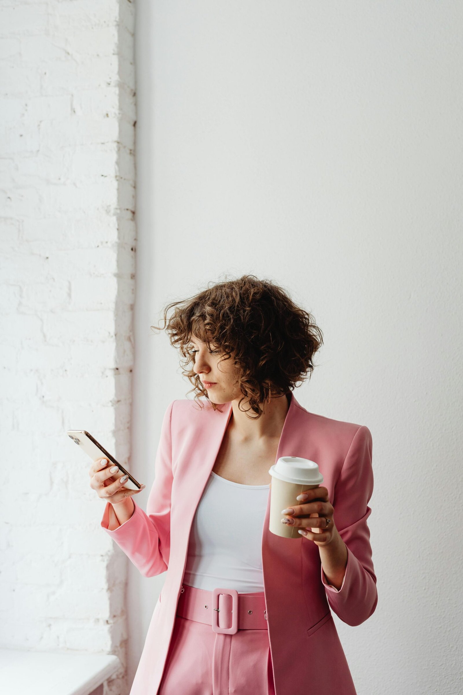 Elegant woman in a pink blazer texting on her smartphone while holding a coffee cup indoors.
